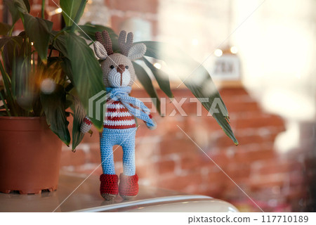 Christmas deer. A Closed up toy amigurumi deer in striped sweater and stylish blue scarf stands near flowerpot. Free copyspace. 117710189