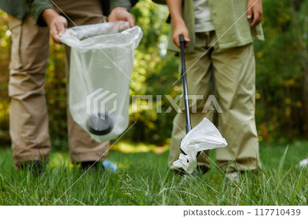 Two individuals picking up litter with trash grabber in wooded park area on sunny day, maintaining cleanliness and contributing to environmental conservation 117710439