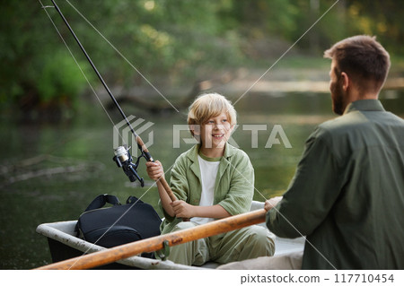 Child and adult enjoying peaceful fishing session on calm lake from small boat while child holding fishing rod having conversation in relaxed setting Child and adult enjoying peaceful fishing session on calm lake from small boat while child holding fishing rod having conversation in relaxed setting 117710454