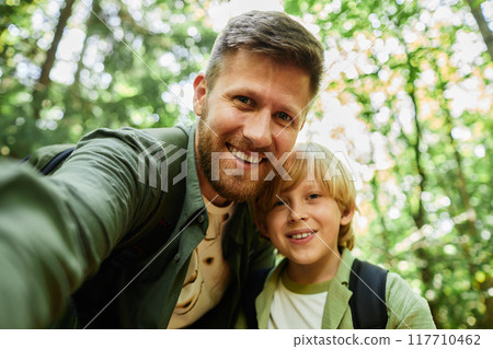 Portrait of father and son smiling for camera while taking selfie in lush green forest showing cheerful moment during outdoor adventure bonding experience 117710462