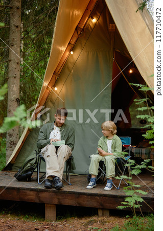 Father and child sharing a moment while sitting near tent in wooded area, enjoying outdoor activity during camping trip in forest 117710472