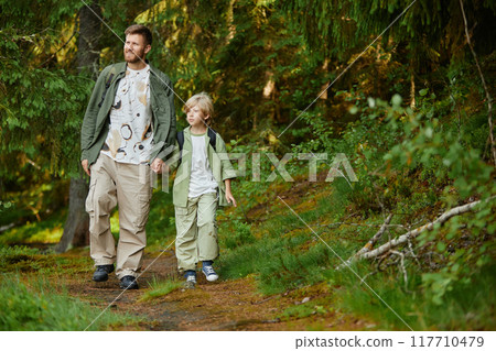 Father and son walking hand in hand along green forest trail on sunny day. Both dressed in casual hiking outfits, enjoying nature and each other's company Father and son walking hand in hand along green forest trail on sunny day. Both dressed in casual hiking outfits, enjoying nature and each other's company 117710479