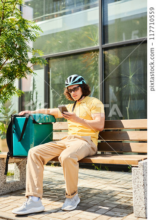 Cyclist with curly hair sitting on bench outside modern building and checking smartphone while resting during delivery work wearing helmet and casual clothing 117710559
