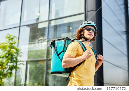 Bicyclist wearing yellow polo shirt and helmet, carrying teal insulated bag for food delivery and standing in front of glass building Bicyclist wearing yellow polo shirt and helmet, carrying teal insulated bag for food delivery and standing in front of glass building 117710572