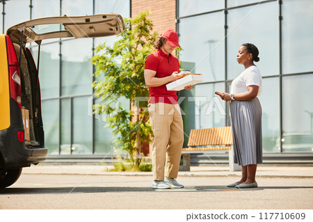 Delivery worker handing over package, with customer receiving it near delivery van. Scene depicts professional service interaction outside a commercial building Delivery worker handing over package, with customer receiving it near delivery van. Scene depicts professional service interaction outside a commercial building 117710609
