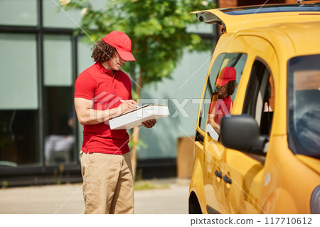 Delivery man in red uniform delivering package while standing next to yellow van in urban area 117710612