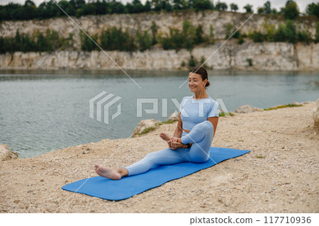 A peaceful woman is practicing yoga techniques by the serene lake during the day 117710936