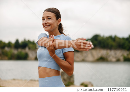 A woman performing stretching exercises outdoors next to the refreshing water body 117710951