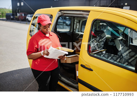 Delivery worker wearing red uniform checking packages beside yellow van in outdoor setting, engrossed in verifying delivery list while holding a package 117710993