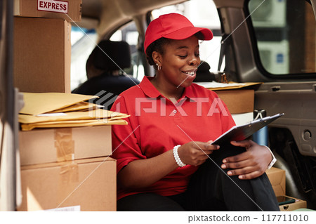 Smiling worker sitting in delivery van organizing and checking packages with clipboard, showcasing organizational skills and efficiency amidst courier tasks Smiling worker sitting in delivery van organizing and checking packages with clipboard, showcasing organizational skills and efficiency amidst courier tasks 117711010