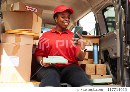 Smiling delivery worker holding package while looking at smartphone in delivery vehicle surrounded by boxes smiling and wearing red uniform Smiling delivery worker holding package while looking at smartphone in delivery vehicle surrounded by boxes smiling and wearing red uniform 117711013