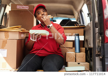 Delivery worker eating during a break while surrounded by packages in van. Wearing uniform and hat and taking moment to relax amidst busy day of deliveries Delivery worker eating during a break while surrounded by packages in van. Wearing uniform and hat and taking moment to relax amidst busy day of deliveries 117711014
