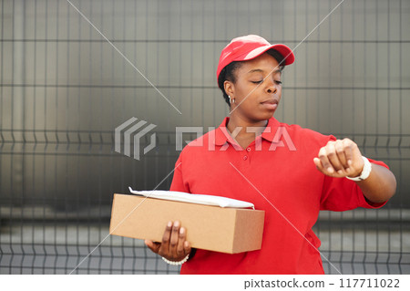 African American woman in red uniform holding a package and glancing at wristwatch. Background features a metallic fence and an industrial setting 117711022