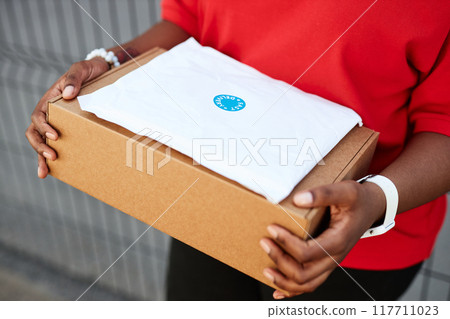 Person holding cardboard package with white label sticker on top wearing red shirt standing outside building Person holding cardboard package with white label sticker on top wearing red shirt standing outside building 117711023