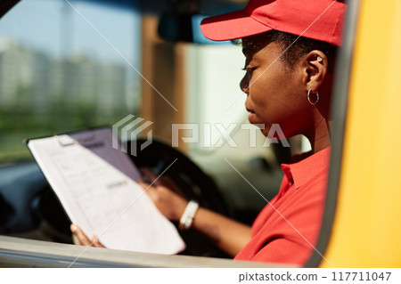 African American woman in red uniform reviewing checklist while sitting in delivery van with focus on task at hand and clipboard in hand. Background shows blurry urban setting African American woman in red uniform reviewing checklist while sitting in delivery van with focus on task at hand and clipboard in hand. Background shows blurry urban setting 117711047