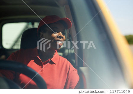 Smiling female delivery worker wearing red cap and shirt while sitting inside a delivery truck and looking outside window 117711049