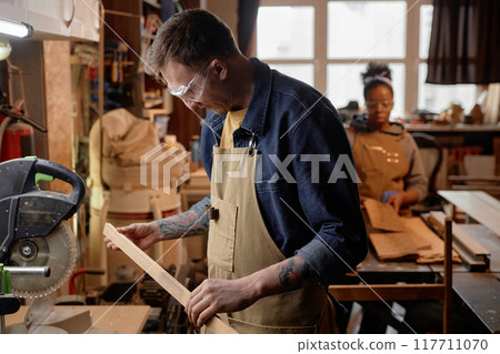 Man working in artisan workshop with focus on wooden piece and tools visible around him. Person in background also engaging in crafting activity in rustic wooden workshop 117711070