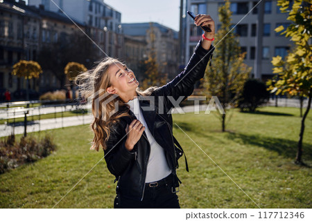 Cheerful beautiful student girl making selfie photo on smart phone on the street, walk outdoors. Cheerful beautiful student girl making selfie photo on smart phone on the street, walk outdoors. 117712346