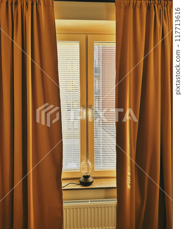 A lamp on the windowsill by the window with blinds and yellow curtains, the setting sun beautifully paints the interior. 117713616