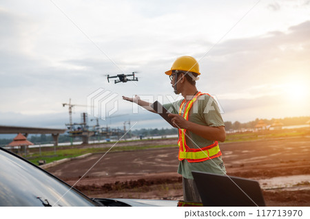 An engineer man in a yellow hard hat is standing next to a car with a drone flying above 117713970