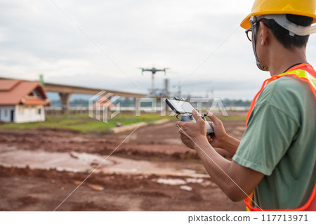 An engineer man in a yellow safety vest is holding a remote control and looking at a drone An engineer man in a yellow safety vest is holding a remote control and looking at a drone 117713971