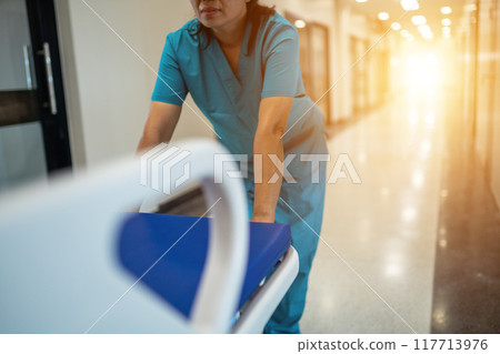 A woman in a blue scrubs is pushing a hospital bed A woman in a blue scrubs is pushing a hospital bed 117713976