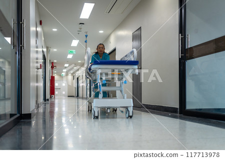 A woman in a green uniform is pushing a hospital bed down a hallway 117713978