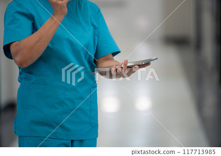 A woman in a blue scrubs is holding a tablet in her hand A woman in a blue scrubs is holding a tablet in her hand 117713985