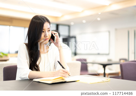 一位女士在筆記本上寫字,同時使用智慧型手機的通話功能通話 一位女士在筆記本上寫字,同時使用智慧型手機的通話功能通話 117714486