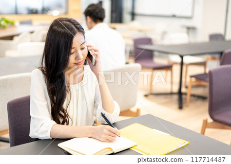 一位女士在筆記本上寫字，同時使用智慧型手機的通話功能進行對話 117714487