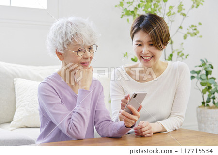 Daughter teaching her mother how to use a smartphone in the living room Daughter teaching her mother how to use a smartphone in the living room 117715534