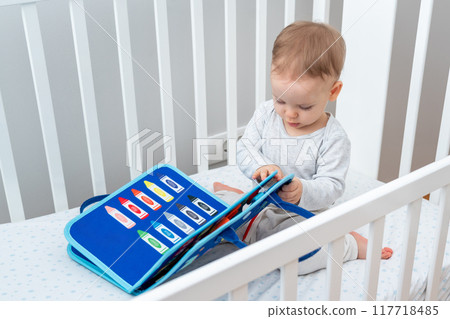 Baby playing with montessori busy book sitting in crib. Concept of smart books and modern toys Baby playing with montessori busy book sitting in crib. Concept of smart books and modern toys 117718485