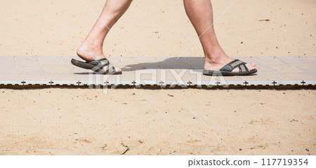 Men's feet in summer slates on the beach sand track. On the yellow sand beach lies on the path and there are feet in the summer slates Men's feet in summer slates on the beach sand track. On the yellow sand beach lies on the path and there are feet in the summer slates 117719354