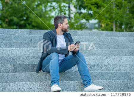 man messaging on phone sitting in the street. man messaging on phone outside. 117719576