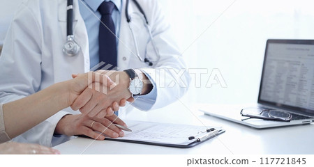 Doctor and a patient are shaking hands. The physician is wearing a white medical coat over a blue shirt. Medical service 117721845