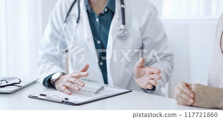 Doctor and a patient. The physician, wearing a white medical coat over a green shirt, is gesturing with his hands during a consultation in the clinic. Medicine 117721860