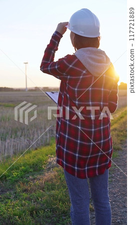 Woman engineer wearing a white protective helmet is taking notes with a clipboard in a field with wind turbines, as the sun sets. Clean energy and engineering concept 117721889