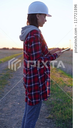 Woman engineer wearing a white protective helmet is taking notes with a clipboard in a field with wind turbines. Clean energy and engineering concept 117721896