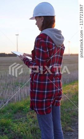 Woman engineer wearing a white protective helmet is taking notes with a clipboard in a field with wind turbines, as the sun sets. Clean energy and engineering concept 117721897