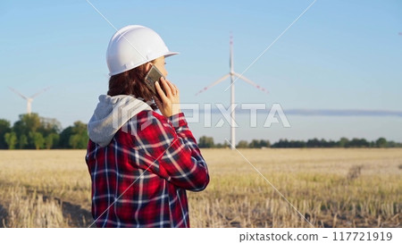 Woman engineer is using a smartphone in a field with wind turbines as the sun sets. Concept of clean energy and engineering audit 117721919