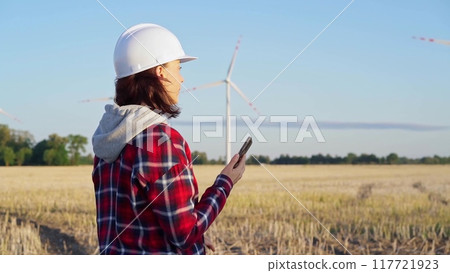 Woman engineer is talking on a smartphone in a field with wind turbines as the sun sets. Concept of clean energy and engineering audit Woman engineer is talking on a smartphone in a field with wind turbines as the sun sets. Concept of clean energy and engineering audit 117721923