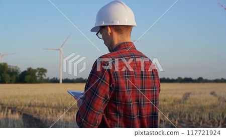 Man engineer at work is wearing a white protective helmet and taking notes with a tablet computer in a field with wind turbines, as the sun sets. Clean energy concept Man engineer at work is wearing a white protective helmet and taking notes with a tablet computer in a field with wind turbines, as the sun sets. Clean energy concept 117721924