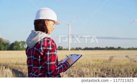Female engineer wearing a white protective helmet is taking notes with a clipboard in a field with wind turbines, as the sun sets. Clean energy concept Female engineer wearing a white protective helmet is taking notes with a clipboard in a field with wind turbines, as the sun sets. Clean energy concept 117721925
