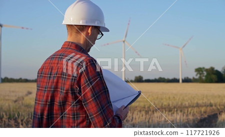 Man engineer, wearing a white protective helmet is taking notes with a clipboard in a field with wind turbines, as the sun sets. Clean energy and engineering concept Man engineer, wearing a white protective helmet is taking notes with a clipboard in a field with wind turbines, as the sun sets. Clean energy and engineering concept 117721926