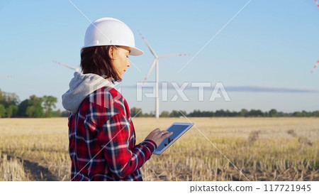 Female engineer wearing a white protective helmet is taking notes with a tablet computer on a field with wind turbines, as the sun sets. Clean energy and engineering 117721945