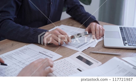 Close up of professional hands using a calculator to conduct financial analysis at the background of a colleague with a magnificent glass. Bookkeeping, finance audit and taxes 117721987