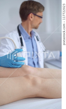 Doctor man with blue protective gloves using ultrasound equipment on the knee of a female patient, analyzing the results on a monitor in the background. Medicine Doctor man with blue protective gloves using ultrasound equipment on the knee of a female patient, analyzing the results on a monitor in the background. Medicine 117722023