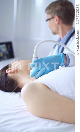 Doctor man wearing blue protective gloves is using ultrasound equipment on the neck of a patient woman, lying for examination. Medicine concept 117722028