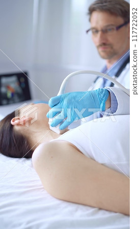 Doctor man wearing blue protective gloves is using ultrasound equipment on the neck of a patient woman, lying for examination. Medicine concept 117722052