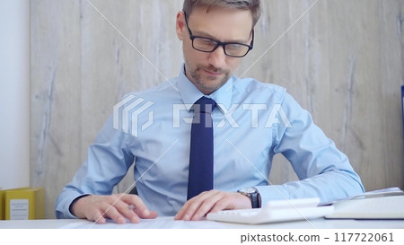 Male accountant with blue t-shirt concentrating while calculating costs and taking notes at his desk. Taxes, audit in business 117722061